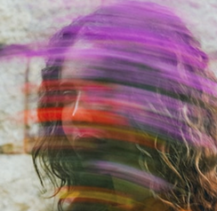 A blurred portrait of a person with long, wavy hair, featuring vibrant streaks of purple, red, and green across the image.