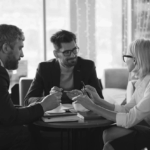 Three professionals engaged in a discussion at a table, with a coffee cup and notepad. The setting is bright, with soft lighting.