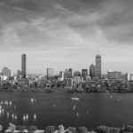 Black and white skyline of Boston with tall buildings, including the Prudential Tower and John Hancock Tower, over the Charles River. Sailboats dot the water.