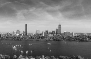 Black and white skyline of Boston with tall buildings, including the Prudential Tower and John Hancock Tower, over the Charles River. Sailboats dot the water.