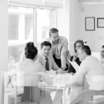 A group of six people in a bright, modern office meeting room, engaged in discussion around a table.