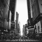 Crowded Times Square with pedestrians crossing a street, surrounded by tall buildings and large advertisements, in black and white.