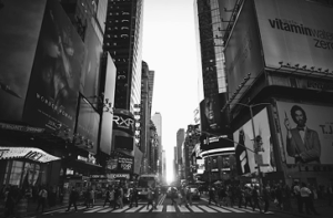 Crowded Times Square with pedestrians crossing a street, surrounded by tall buildings and large advertisements, in black and white.