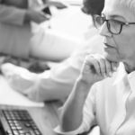 A woman with short gray hair and glasses sits at a desk, thoughtfully resting her chin on her hand, focused on a computer.