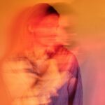 A blurred portrait of a woman with light brown hair, wearing a striped shirt against a warm orange background, conveying motion.