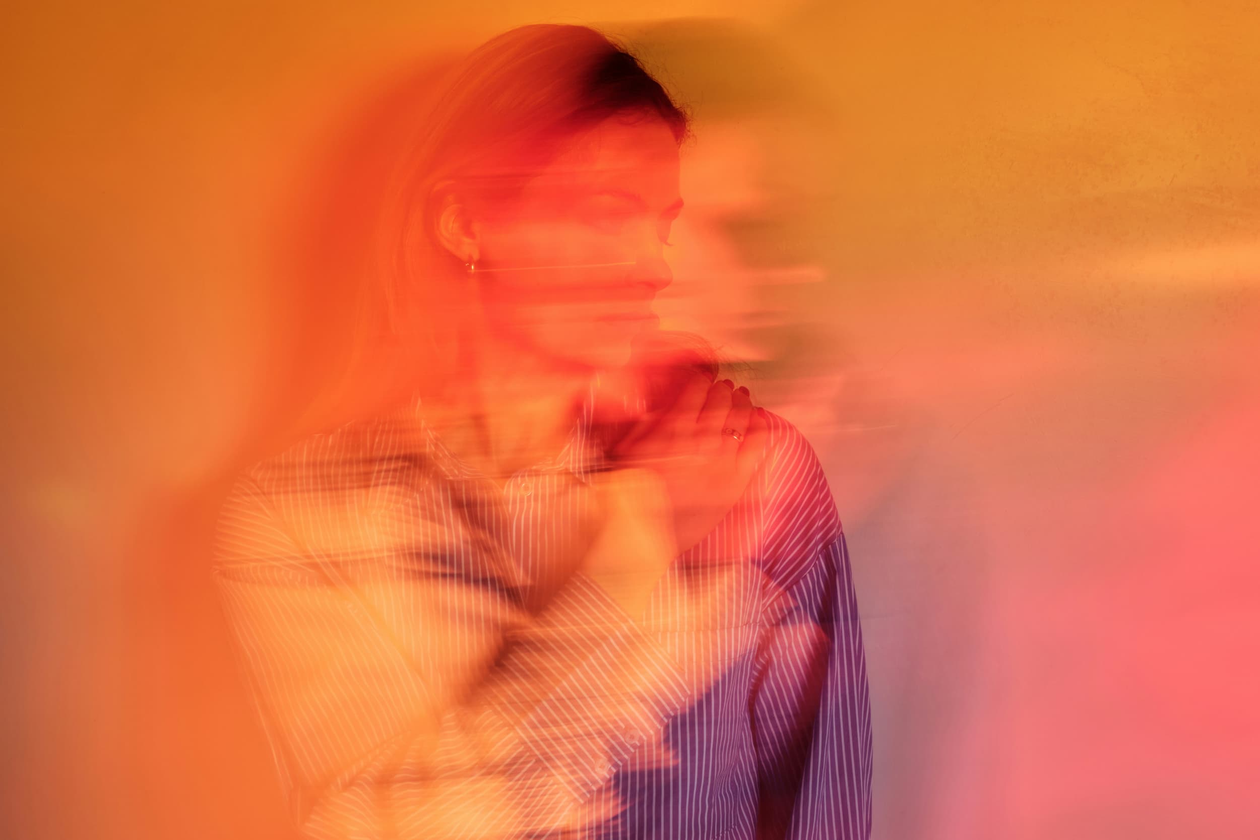 A blurred portrait of a woman with light brown hair, wearing a striped shirt against a warm orange background, conveying motion.