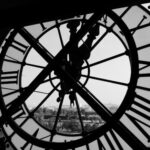 Large clock face with Roman numerals and hands, set against a view of a cityscape in black and white.