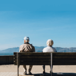 Elderly couple sitting on a bench, facing the ocean and mountains under a clear blue sky.